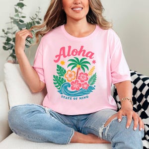 A woman wearing a pink t-shirt with a tropical design and the text "Aloha State of Mind" is sitting on a couch.