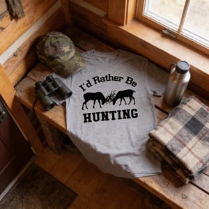 a gray t-shirt with the text "I'd Rather Be Hunting" printed on it, along with a camo hat and binoculars. The t-shirt is placed on a wooden bench, and there is a bottle and a blanket nearby.