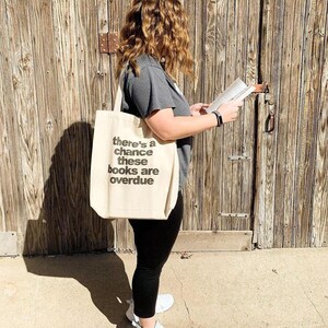 A woman standing in front of a wooden door, holding a book and wearing a tote bag with text on it.