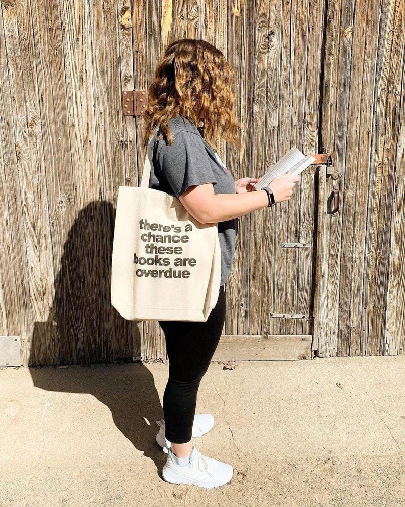 A woman standing in front of a wooden door, holding a book and wearing a tote bag with text on it.