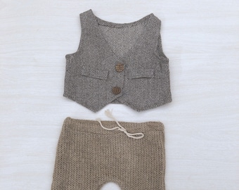 Könnte beinhalten: Ein zweiteiliges Baby-Outfit. Das Oberteil ist eine graue Weste mit Rautenmuster und Holzknöpfen. Die Hose ist eine gestrickte braune Hose mit Kordelzug. Das Outfit ist für ein Neugeborenes konzipiert.