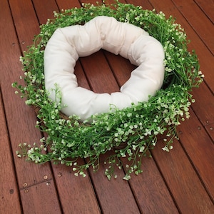 Puede incluir: Un cojín blanco redondo acolchado con un agujero en el centro, rodeado de una corona de enredaderas verdes con pequeñas flores blancas.