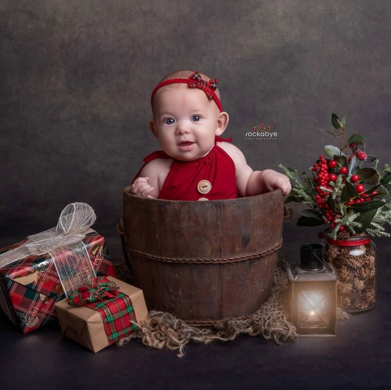 May include: A baby wearing a red and white checked headband and a red sleeveless onesie sits in a wooden bucket. The baby is surrounded by Christmas-themed gifts and decorations.