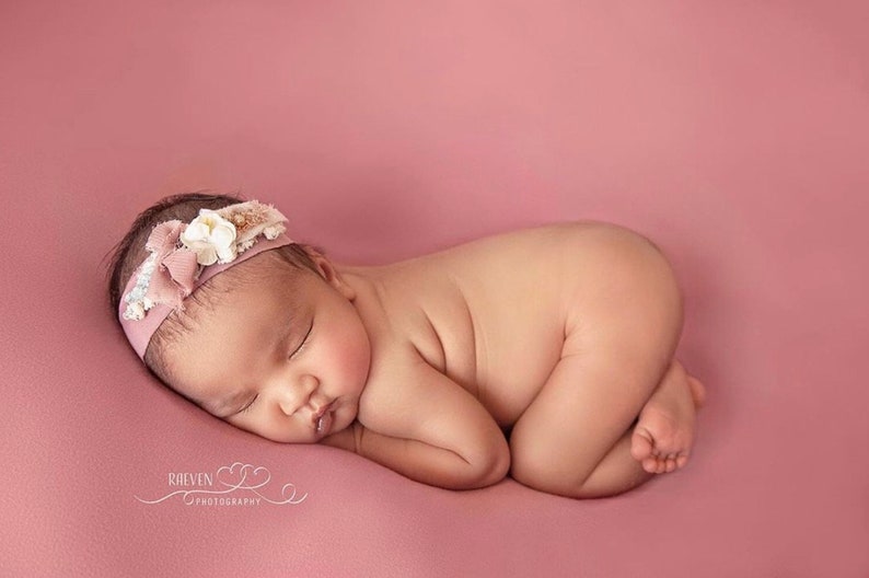Pu&ograve; includere: Una neonata dorme su uno sfondo rosa. Indossa una fascia rosa con fiori bianchi e pizzo.