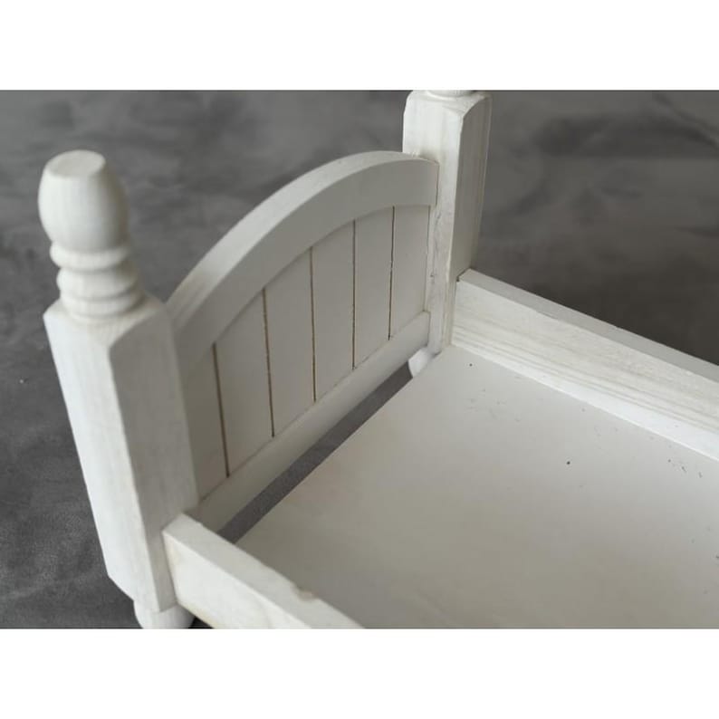 Puede incluir: Un peque&ntilde;o marco de cama de madera blanca con una cabecera con listones verticales y postes redondeados. El marco de la cama est&aacute; pintado de blanco y parece ser un mueble en miniatura o del tama&ntilde;o de una mu&ntilde;eca.