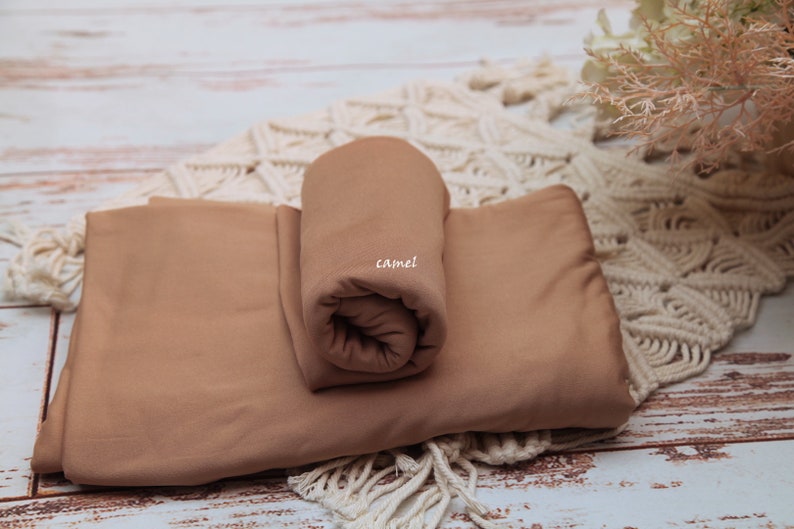 Pu&ograve; includere: Due pezzi di tessuto color cammello, uno arrotolato e l'altro piatto, su una superficie di uncinetto bianca. Il tessuto arrotolato ha la scritta "camel".