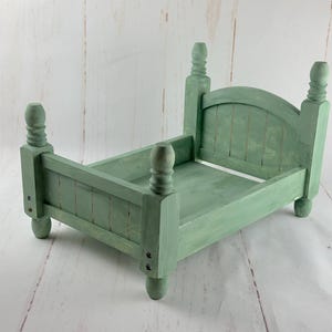 Puede incluir: Una peque&ntilde;a estructura de cama de madera verde salvia con cabecero y piecero. La cama tiene cuatro postes decorativos y est&aacute; pintada al estilo envejecido. La estructura de la cama est&aacute; dise&ntilde;ada para accesorios de fotograf&iacute;a.