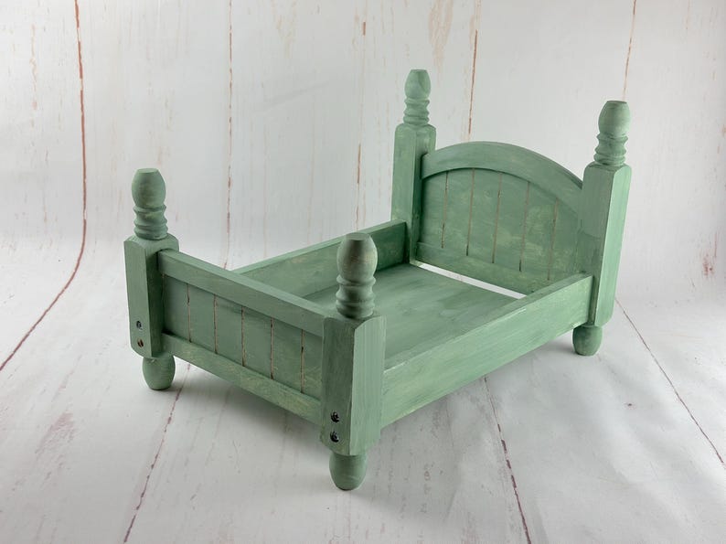 Puede incluir: Una peque&ntilde;a estructura de cama de madera verde salvia con cabecero y piecero. La cama tiene cuatro postes decorativos y est&aacute; pintada al estilo envejecido. La estructura de la cama est&aacute; dise&ntilde;ada para accesorios de fotograf&iacute;a.