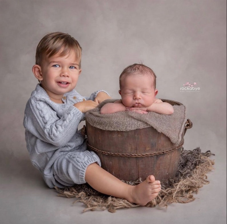 May include: A toddler wearing a light blue knitted onesie sits beside a wooden bucket containing a newborn baby. The baby is wrapped in a beige blanket and is asleep. The bucket is placed on a bed of brown and grey straw.