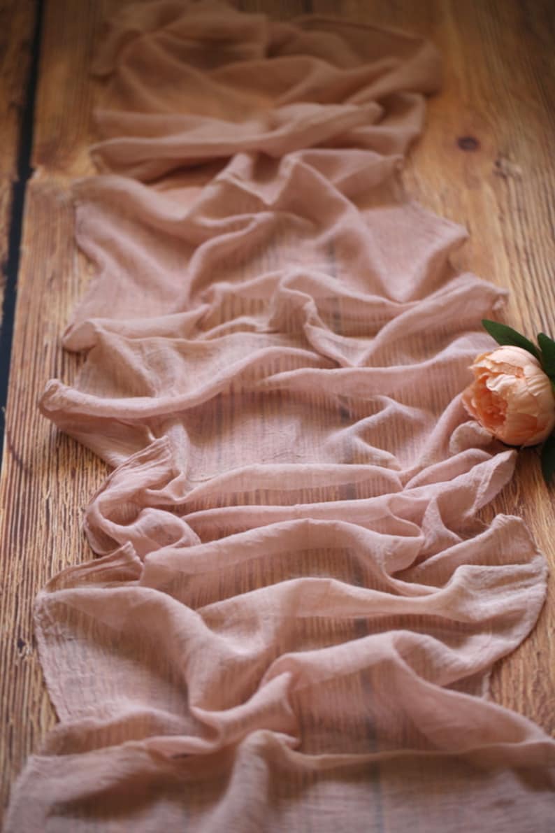 Pu&ograve; includere: Un runner da tavolo in tessuto rosa chiaro trasparente con una texture morbida e fluida. Il runner &egrave; drappeggiato su una superficie in legno.