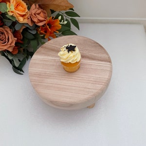 Puede incluir: Un soporte para pastel de madera con un solo cupcake en la parte superior. El cupcake est&aacute; cubierto con glaseado blanco y una estrella negra.