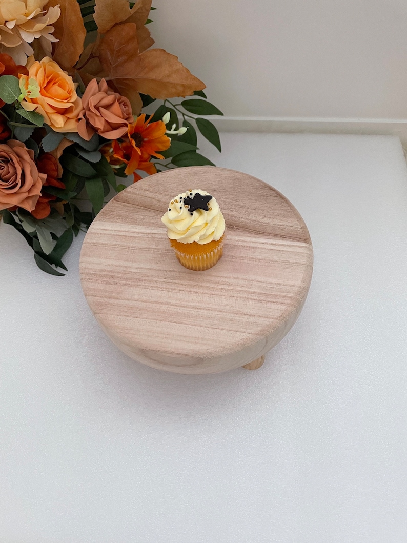 Puede incluir: Un soporte para pastel de madera con un solo cupcake en la parte superior. El cupcake est&aacute; cubierto con glaseado blanco y una estrella negra.