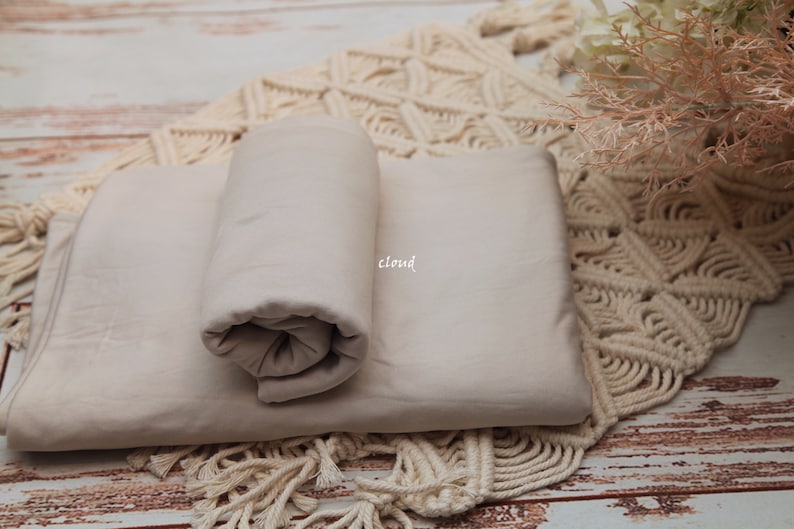 Pu&ograve; includere: Uno sfondo in tessuto beige per la fotografia, arrotolato e posizionato su un tappeto all'uncinetto color crema. Il tessuto &egrave; liscio e ha una lucentezza sottile.