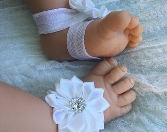 May include: White baby foot jewellery with a white satin flower and a rhinestone centre. The jewellery is tied with a white ribbon.