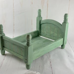 Puede incluir: Una peque&ntilde;a cama de madera verde salvia con cuatro postes decorativos y una cabecera. La cama est&aacute; pintada con un acabado envejecido, lo que le da un aspecto vintage. El marco de la cama est&aacute; dise&ntilde;ado para accesorios de fotograf&iacute;a.