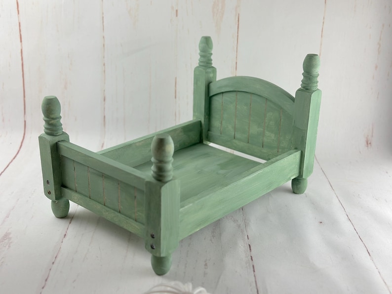Puede incluir: Una peque&ntilde;a cama de madera verde salvia con cuatro postes decorativos y una cabecera. La cama est&aacute; pintada con un acabado envejecido, lo que le da un aspecto vintage. El marco de la cama est&aacute; dise&ntilde;ado para accesorios de fotograf&iacute;a.