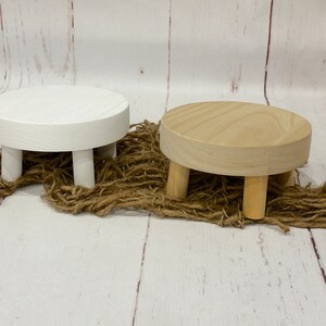 Puede incluir: Dos soportes de pedestal redondos de madera con cuatro patas. Un soporte es blanco y el otro es de madera natural.