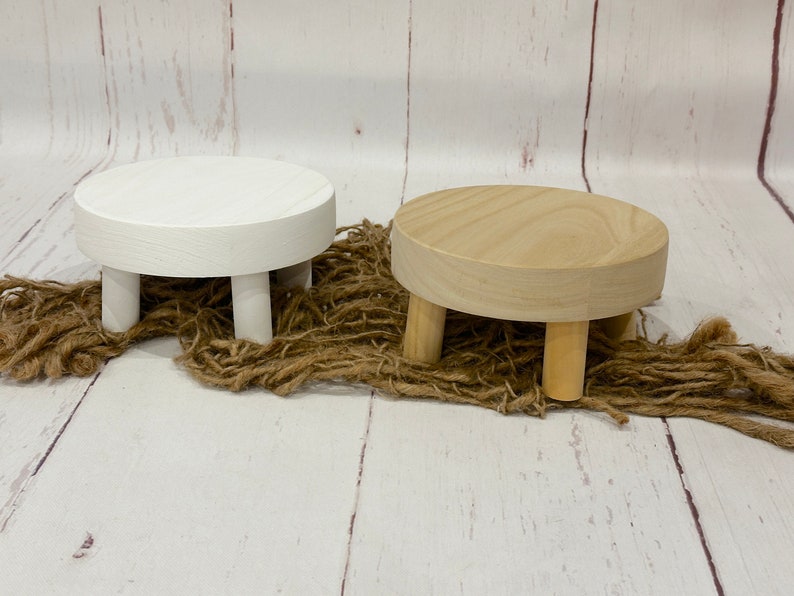 Puede incluir: Dos soportes de pedestal redondos de madera con cuatro patas. Un soporte es blanco y el otro es de madera natural.