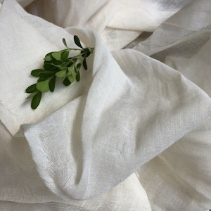 May include: A white, sheer fabric with a delicate texture. The fabric is folded and draped, creating soft folds and wrinkles. A small sprig of green leaves is placed on top of the fabric.