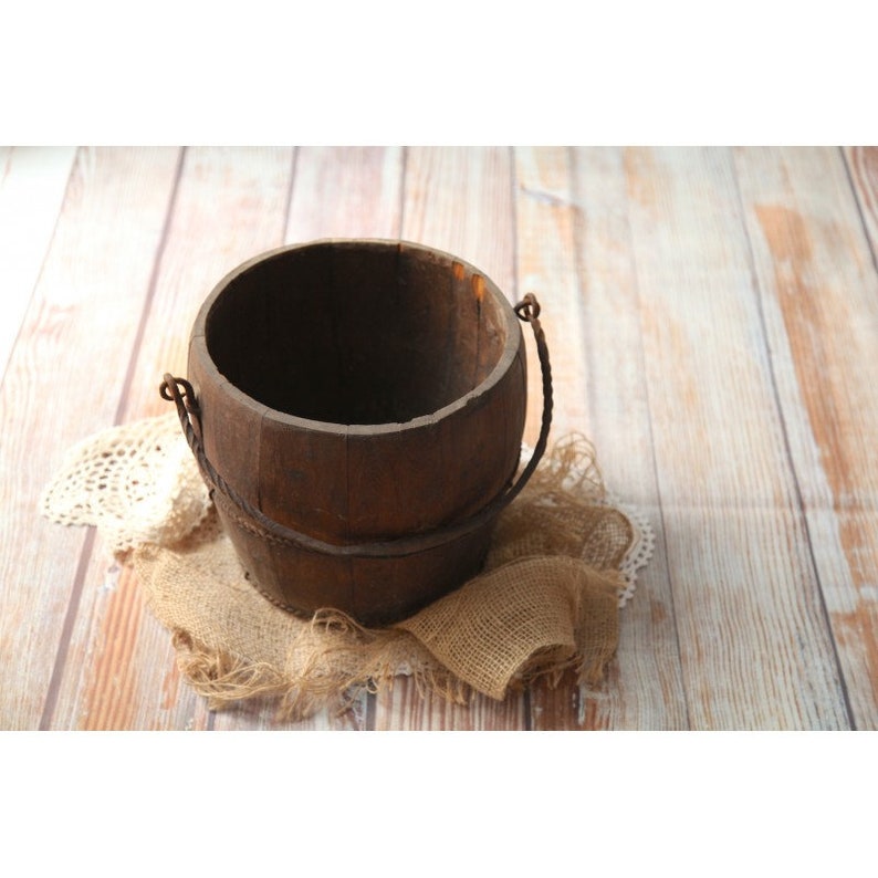 May include: A rustic wooden bucket with a metal handle. The bucket is brown and has a worn, weathered look. It is sitting on a hessian cloth and a white lace doily.