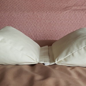 May include: A white, wedge-shaped pillow with a zipper in the center. The pillow is made of a soft, fabric material and is designed to provide support and comfort.