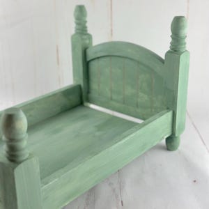 Puede incluir: Una peque&ntilde;a estructura de cama de madera verde salvia con cabecero y piecero. La cama tiene cuatro postes decorativos y un cabecero de listones. La estructura de la cama mide aproximadamente 25 cm de largo y 15 cm de ancho, con un acabado de pintura envejecida.