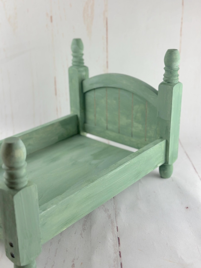Puede incluir: Una peque&ntilde;a estructura de cama de madera verde salvia con cabecero y piecero. La cama tiene cuatro postes decorativos y un cabecero de listones. La estructura de la cama mide aproximadamente 25 cm de largo y 15 cm de ancho, con un acabado de pintura envejecida.