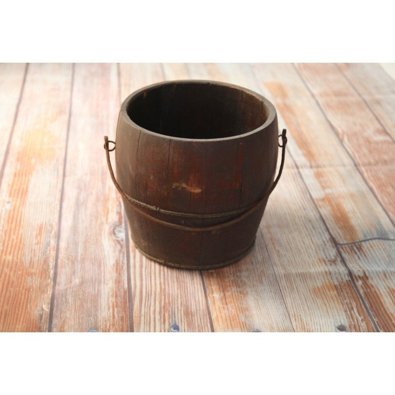 May include: A rustic wooden bucket with a metal handle. The bucket is brown and has a worn, weathered look.