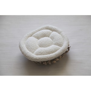 May include: White oval-shaped newborn photography prop with a padded, quilted surface. The prop is made of soft, fluffy material and is designed to provide a comfortable and safe space for a newborn baby.
