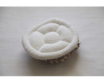 May include: White oval-shaped newborn photography prop with a padded, quilted surface. The prop is made of soft, fluffy material and is designed to provide a comfortable and safe space for a newborn baby.