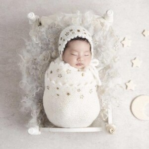 Puede incluir: Un reci&eacute;n nacido dormido envuelto en una manta y gorro de punto blanco, adornado con estrellas doradas. El beb&eacute; descansa en una peque&ntilde;a cama blanca con una manta blanca y esponjosa, rodeado de decoraciones de estrellas y luna creciente.