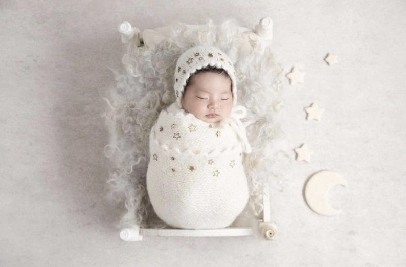 Puede incluir: Un reci&eacute;n nacido dormido envuelto en una manta y gorro de punto blanco, adornado con estrellas doradas. El beb&eacute; descansa en una peque&ntilde;a cama blanca con una manta blanca y esponjosa, rodeado de decoraciones de estrellas y luna creciente.