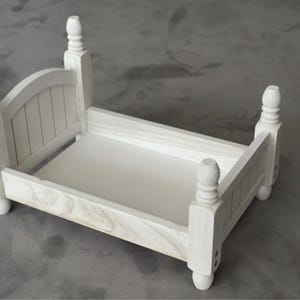 Puede incluir: Un peque&ntilde;o marco de cama de madera blanca con cabecero y piecero. La cama tiene cuatro postes decorativos y una base rectangular. El marco de la cama est&aacute; dise&ntilde;ado para accesorios de fotograf&iacute;a o exhibiciones en miniatura.