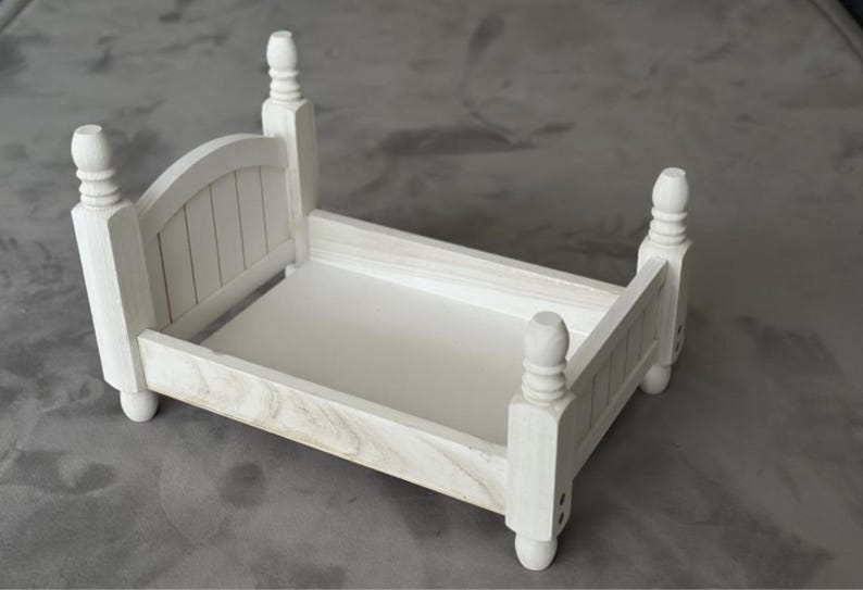 Puede incluir: Un peque&ntilde;o marco de cama de madera blanca con cabecero y piecero. La cama tiene cuatro postes decorativos y una base rectangular. El marco de la cama est&aacute; dise&ntilde;ado para accesorios de fotograf&iacute;a o exhibiciones en miniatura.