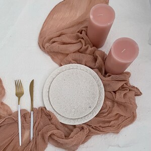 Pu&ograve; includere: Un piatto in ceramica bianca con un motivo floreale, due candele rosa e una forchetta e un coltello dorati su uno sfondo di tessuto marrone chiaro.