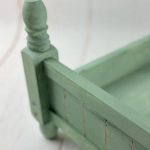 Puede incluir: Un peque&ntilde;o marco de cama de madera verde salvia con una cabecera decorativa. El marco de la cama tiene forma rectangular con listones verticales en el lateral y un pie redondeado. La cabecera presenta un dise&ntilde;o de poste torneado.