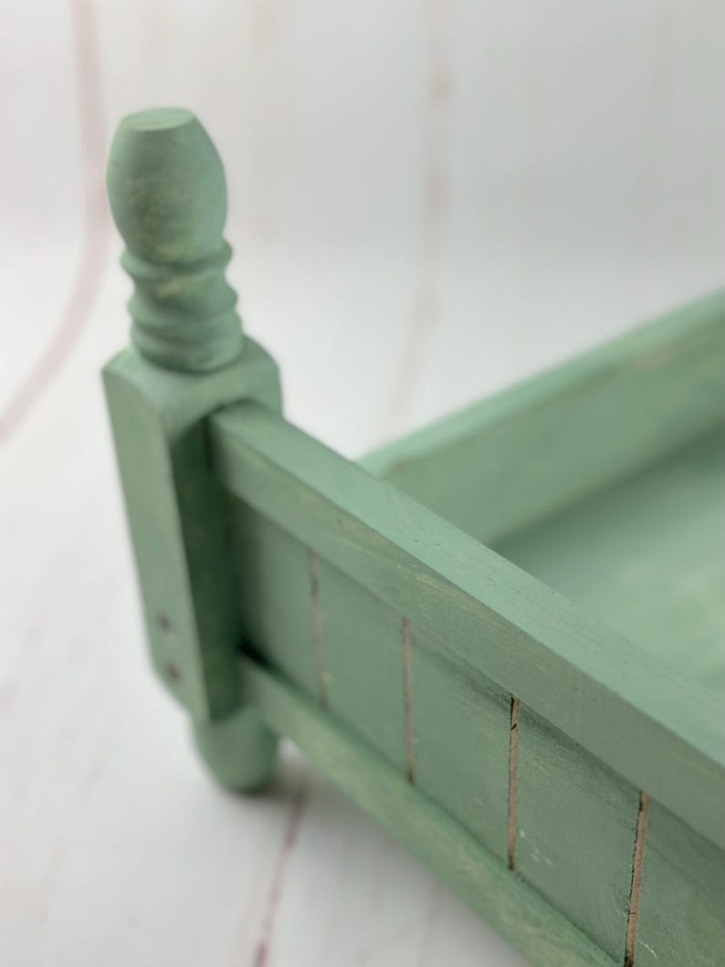 Puede incluir: Un peque&ntilde;o marco de cama de madera verde salvia con una cabecera decorativa. El marco de la cama tiene forma rectangular con listones verticales en el lateral y un pie redondeado. La cabecera presenta un dise&ntilde;o de poste torneado.