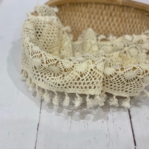 Puede incluir: Una cesta tejida con un forro y ribete de encaje color crema. El encaje tiene un diseño calado delicado con pequeñas borlas a lo largo del borde. La cesta está sobre una superficie de madera blanca.