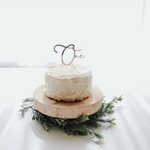 Puede incluir: Un pastel blanco glaseado con un adorno dorado de "Uno" en un soporte de pastel de madera. El pastel est&aacute; rodeado de follaje verde.