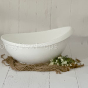 May include: A white, oval-shaped prop bowl for newborn photography. The bowl is set on a bed of twine and greenery against a white wooden backdrop. The text "HoneyDewProps" is visible on the side of the bowl.