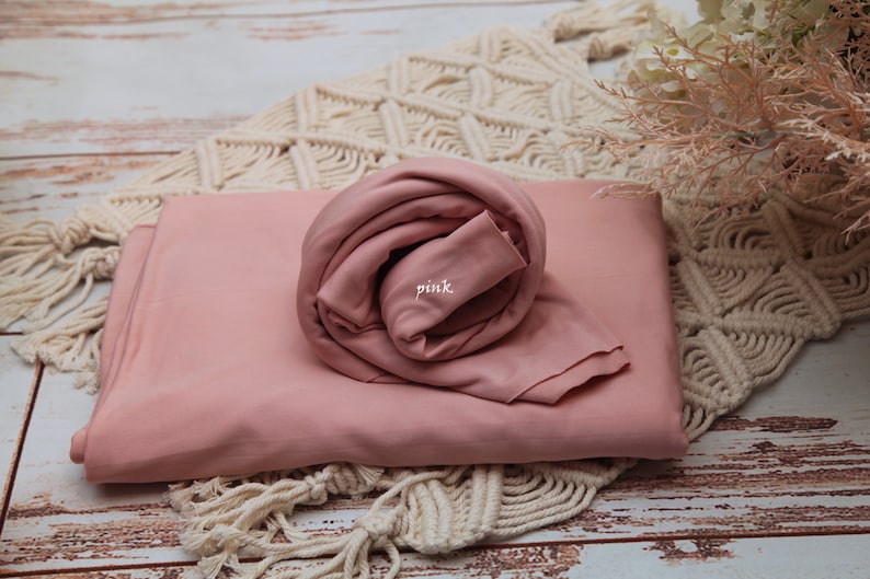 Pu&ograve; includere: Una copertina in tessuto rosa per fasciare i neonati, arrotolata su una superficie di uncinetto bianca. Il tessuto &egrave; morbido e liscio, e la copertina &egrave; perfetta per la fotografia di neonati.