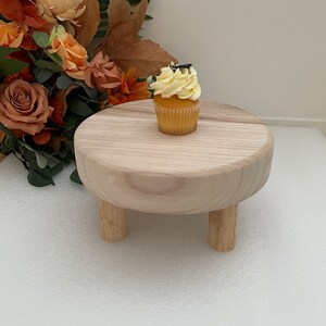 Puede incluir: Un peque&ntilde;o soporte para pasteles de madera con tres patas. El soporte est&aacute; hecho de madera natural y tiene una parte superior redonda. Un cupcake amarillo con chispas negras est&aacute; en la parte superior del soporte.