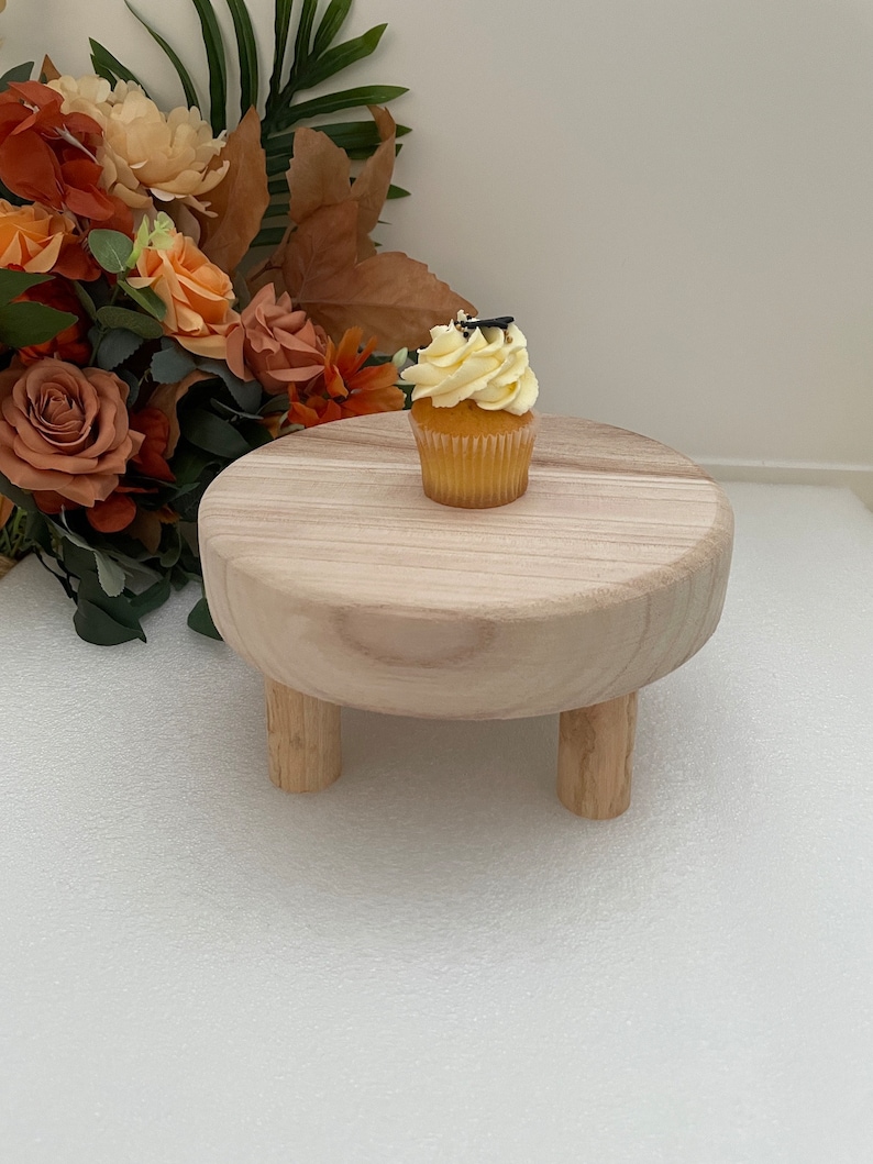 Puede incluir: Un peque&ntilde;o soporte para pasteles de madera con tres patas. El soporte est&aacute; hecho de madera natural y tiene una parte superior redonda. Un cupcake amarillo con chispas negras est&aacute; en la parte superior del soporte.