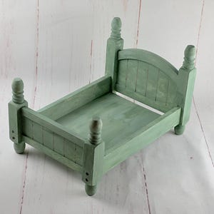 Puede incluir: Una peque&ntilde;a cama de madera verde salvia con cabecero y piecero. La cama tiene cuatro postes decorativos y est&aacute; dise&ntilde;ada para accesorios de fotograf&iacute;a. El marco de la cama mide aproximadamente 30 cm de largo y 20 cm de ancho.