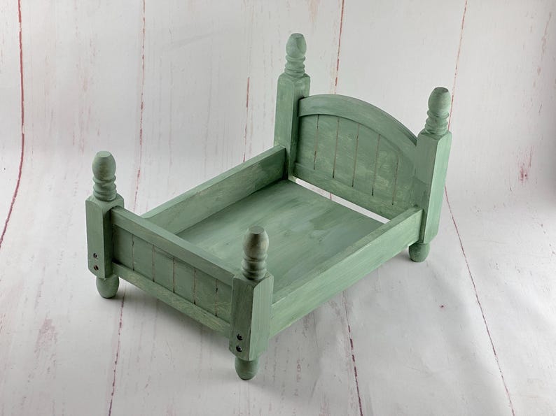 Puede incluir: Una peque&ntilde;a cama de madera verde salvia con cabecero y piecero. La cama tiene cuatro postes decorativos y est&aacute; dise&ntilde;ada para accesorios de fotograf&iacute;a. El marco de la cama mide aproximadamente 30 cm de largo y 20 cm de ancho.