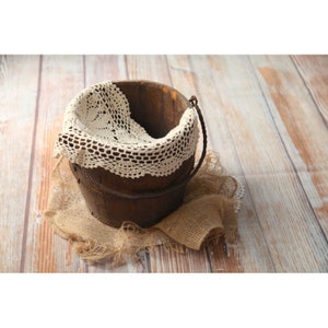 May include: A rustic wooden bucket with a metal handle, lined with a white crocheted lace doily. The bucket is sitting on a hessian fabric background.
