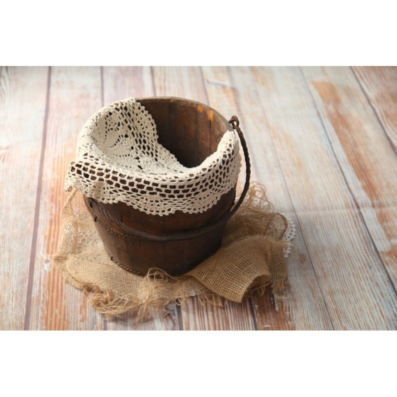 May include: A rustic wooden bucket with a metal handle, lined with a white crocheted lace doily. The bucket is sitting on a hessian fabric background.