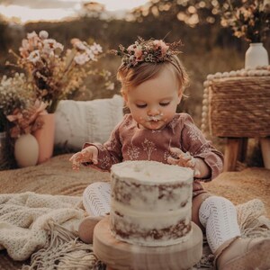 Puede incluir: Una ni&ntilde;a peque&ntilde;a con una corona de flores y un vestido rosa est&aacute; sentada en un taburete de madera y comiendo un pastel blanco glaseado. El pastel est&aacute; en un plato de madera. La ni&ntilde;a est&aacute; rodeada de una manta blanca y un ramo de flores secas.