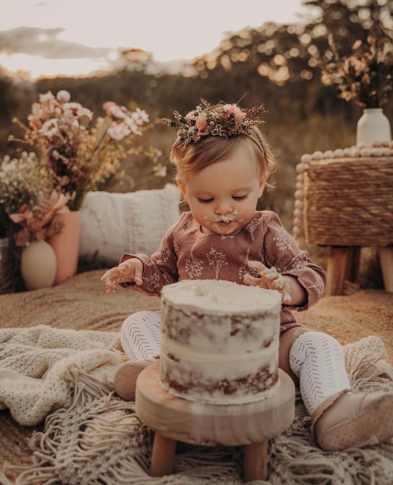 Puede incluir: Una ni&ntilde;a peque&ntilde;a con una corona de flores y un vestido rosa est&aacute; sentada en un taburete de madera y comiendo un pastel blanco glaseado. El pastel est&aacute; en un plato de madera. La ni&ntilde;a est&aacute; rodeada de una manta blanca y un ramo de flores secas.