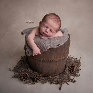 May include: A newborn baby sleeping soundly in a wooden bucket lined with a soft, grey blanket. The bucket is surrounded by a brown, textured fabric and pine cones.